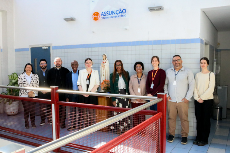 Membros da Secretaria Municipal de Direitos Humanos e Cidadania de São Paulo, ao lado da Reitora e representantes do Centro Universitário Assunção, posam para foto durante a visita institucional. O grupo está em um ambiente interno, com paredes de azulejos claros e o logotipo da instituição ao fundo.