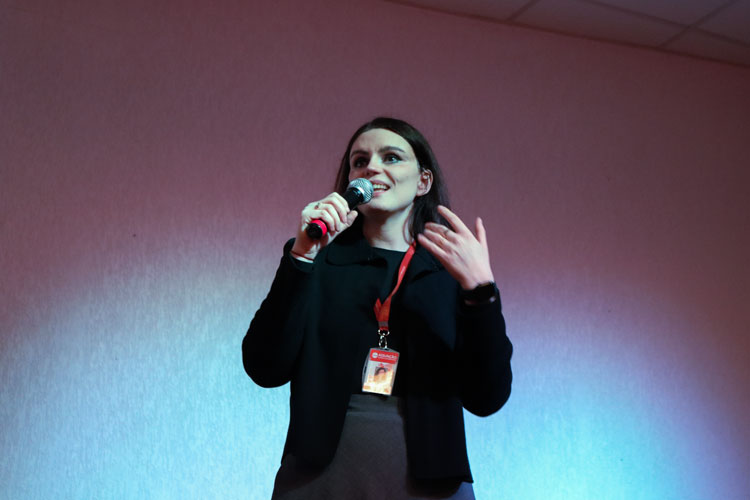 Professora Karen Ambra segura um microfone e gesticula durante sua fala no evento, sob iluminação azul e vermelha no palco.