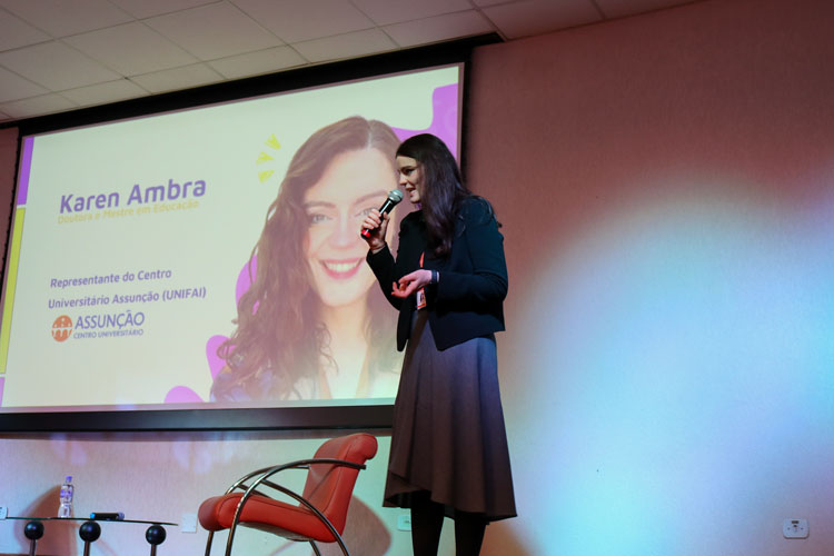 Professora Karen Ambra sorri e fala ao microfone no palco do auditório, com tela ao fundo exibindo sua imagem e nome.