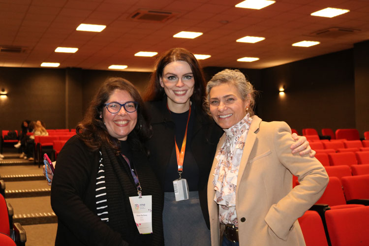 Professora Karen Ambra entre duas mulheres sorridentes em um auditório com cadeiras vermelhas, posando para a foto durante o evento.