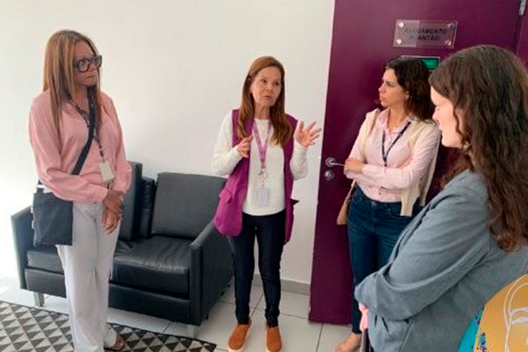 Lucia Brugnera, gestora da Casa da Mulher Brasileira, apresenta informações sobre o funcionamento do local para a Prof.ª Dr.ª Karen Ambra, Dr.ª Regina Célia da Silveira Santana e Dr.ª Stella Verzolla. O grupo está em um ambiente interno, próximo a uma sala de acolhimento.