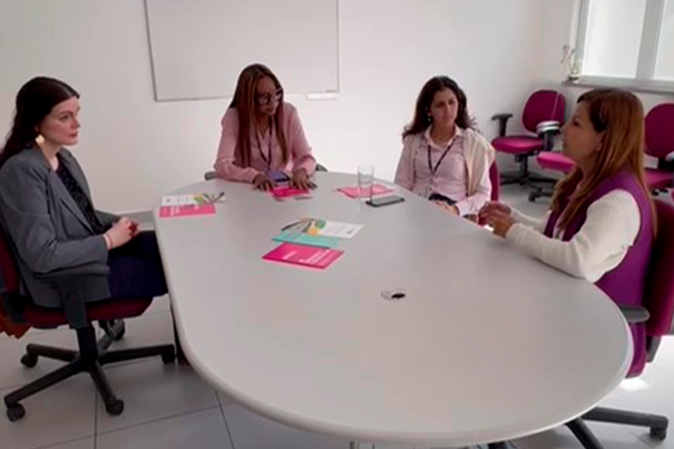 A Prof.ª Dr.ª Karen Ambra, Dr.ª Regina Célia da Silveira Santana, Dr.ª Stella Verzolla e Lucia Brugnera participam de uma reunião ao redor de uma mesa oval, com materiais informativos sobre os serviços da Casa da Mulher Brasileira. O encontro ocorre em uma sala de reuniões, com cadeiras e janelas ao fundo.