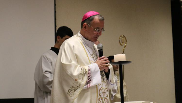 Dom Carlos Lema, de vestes litúrgicas brancas e solidéu rosa, segura um microfone enquanto celebra a missa, com um acólito ao fundo.
