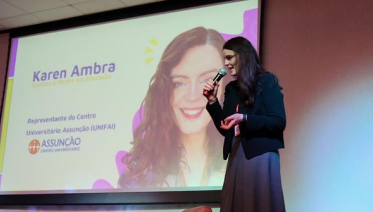 Professora Karen Ambra sorri e fala ao microfone no palco do auditório, com tela ao fundo exibindo sua imagem e nome.
